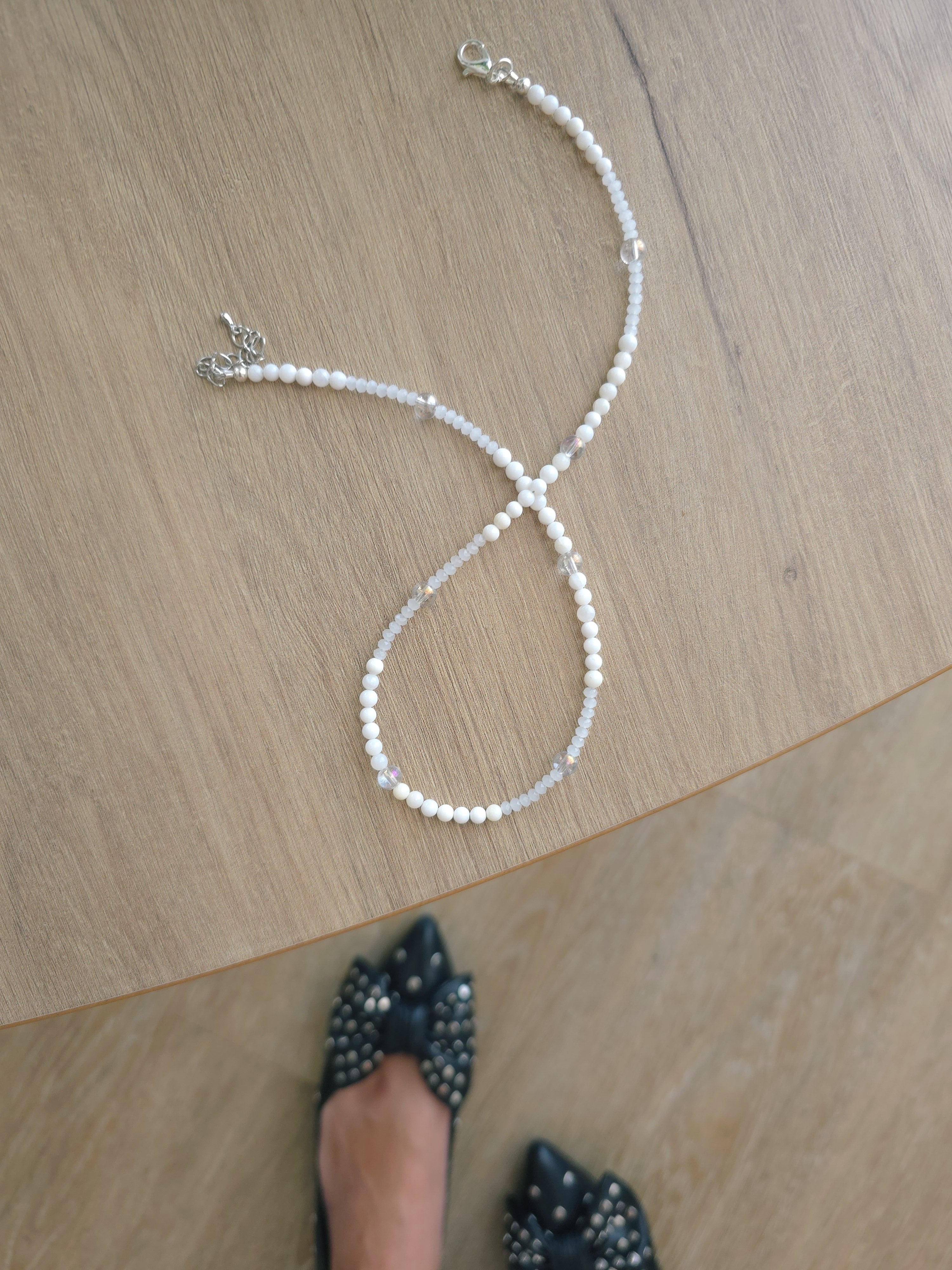 Snow white beaded layering necklace placed on beige coffee table Also black fancy shoes in photo